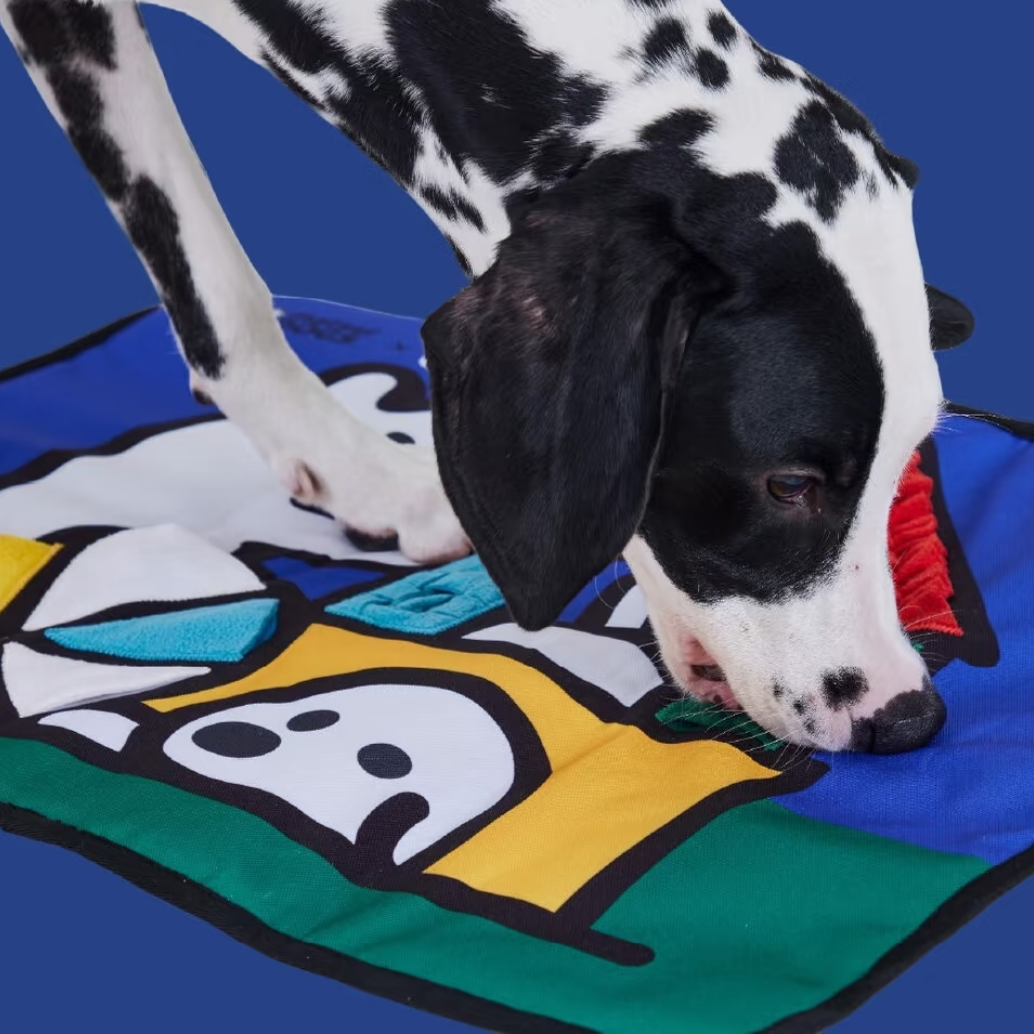 Showing a dog play mat with a black-and-white dog sniffing it on a blue background. The mat has cartoon-style graphics in yellow, green, white, and red, with text overlays describing benefits like 'Sniff for ten minutes, Equal to a dog for an hour.